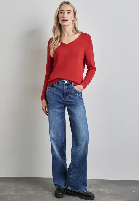 Frau mit rotem Pullover und blauer Jeans steht vor grauer Wand.